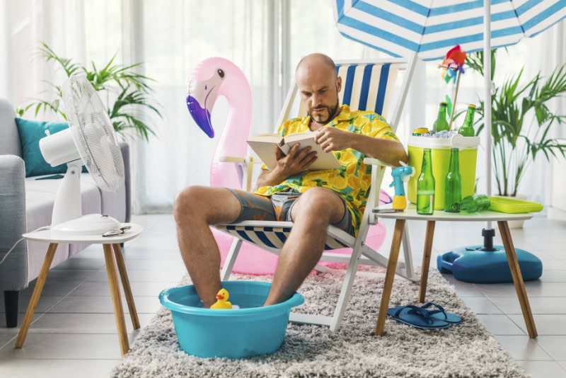 Man spending his summer vacations at home, he is sitting on a deckchair in the living room and reading a book.